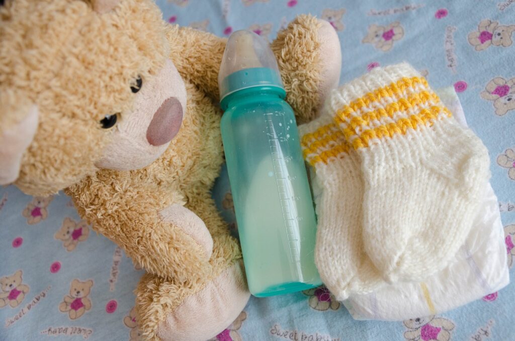 A tan stuffed bear, aqua colored baby bottle, and white socks with yellow stripes.