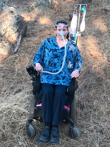 COVID-19 Spokane. Image of Keirsten in her wheelchair, with an IV pole holding feeding tube supplies, and ventilator hosing connected to her face.