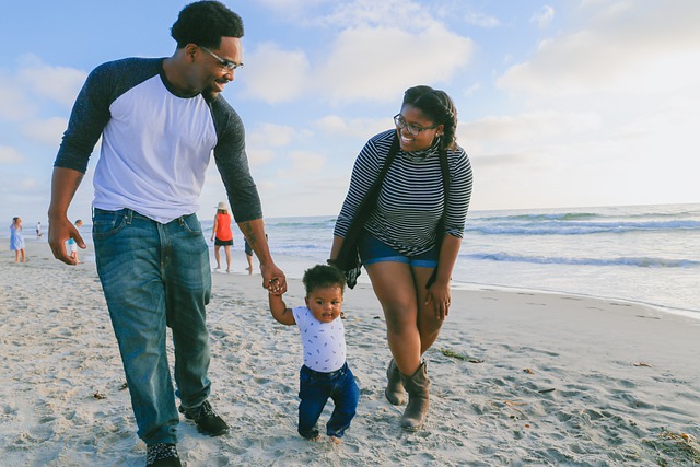 A mother and father holding hands with their toddler, and walking along the sand on a beach.